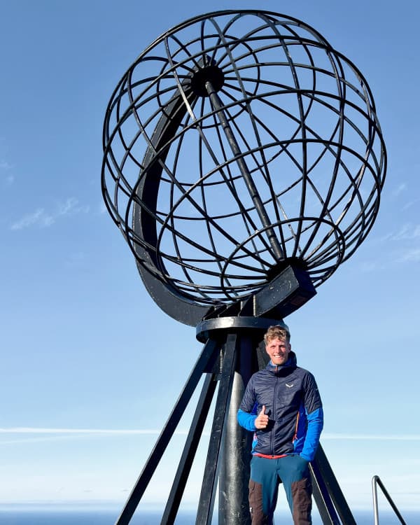 Chris (@van4life__) standing in front of the iconic globe monument at the North Cape, the northernmost point in Europe. Smiling and giving a thumbs up, Chris marks a key moment of his Arctic Circle adventure under clear blue skies. This photo highlights the significance of reaching this landmark during his vanlife journey.