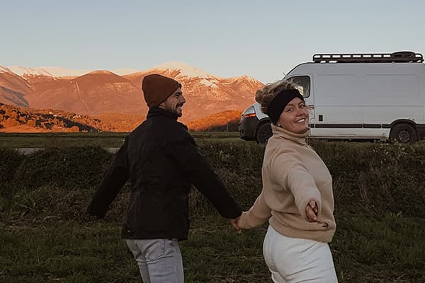 A young couple, @irisandmax, holding hands and smiling in front of their converted Renault Master van. The scenic backdrop features snow-capped mountains and a golden sunset, embodying the essence of vanlife and outdoor exploration.