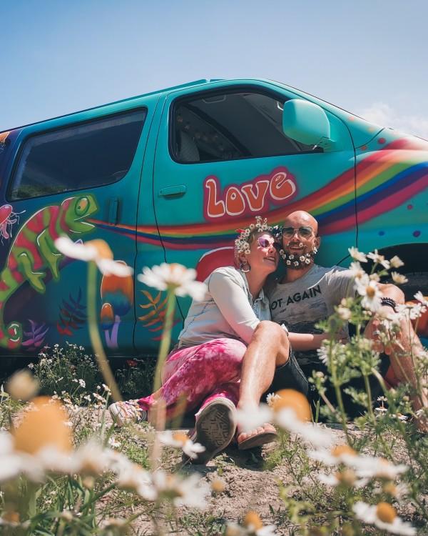 Joriën and Sandra from travelwildtobefree, sitting amidst wildflowers, smiling and enjoying nature in front of their colorful, hippie-themed Toyota HiAce 4x4 camper van.