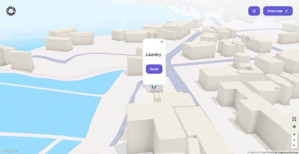 A 3D interactive city map from Vanlifezone, featuring a simplified representation of an urban layout with block-like buildings and blue water. A pop-up on the map highlights a 'Laundry' location with a 'Route' button to guide users to the service. The interface includes a 'Close map' button and map navigation tools to the right, as well as a Mapbox and OpenStreetMap attribution in the lower right corner, indicating the map's source.