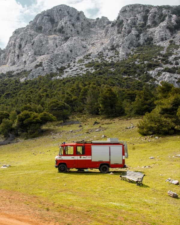 A red firetruck, @hummel_die_feuerwehr, converted into a camper parked in a grassy field at the base of a rocky mountain, showcasing the vanlife experience in a remote and rugged natural setting on Crete’s south coast.