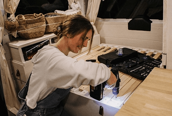 A woman (@itsme.jenni) working inside her self-built camper van, using a power drill to assemble furniture. The van interior features cozy decor and organized tools, reflecting her journey of transforming a VW T5 into a mobile home.