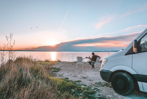 Man sitting by a serene lakeside at sunset, using a guitar, next to a parked white van with the door open. The scene captures a peaceful evening with a vibrant sky reflected in the water, wild grass in the foreground, and birds flying overhead.