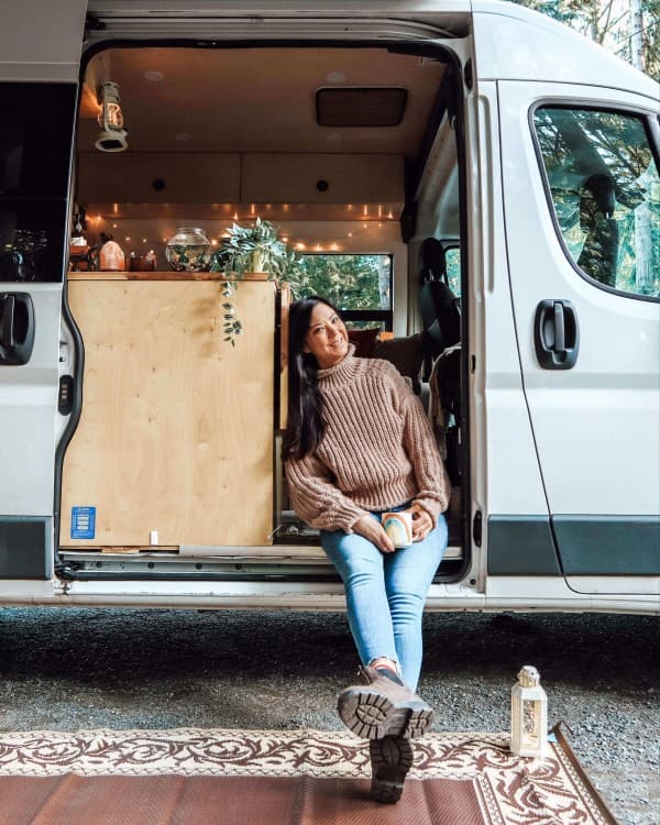 Emily Jay, a solo female traveler, sitting in the open doorway of her white RAM van, which is set up for vanlife. The van interior is decorated with string lights and plants, creating a cozy atmosphere. Emily is holding a colorful mug, wearing a brown sweater and jeans, and is parked in a wooded area, capturing the essence of her vanlife experience in Canada.