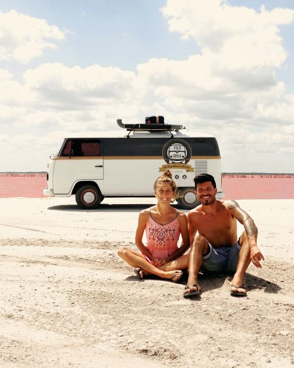 Belu and Lucho sitting in front of their Volkswagen Kombi named Blanquita, parked by the pink waters of Las Coloradas, embracing their slow travel lifestyle and sharing unique experiences.