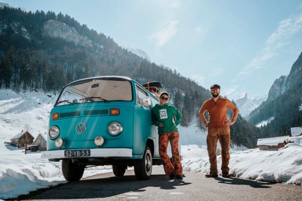 Milene and Yuri standing beside their turquoise Volkswagen T2 van, Alexine, on a snowy mountain road. The backdrop features snow-covered mountains and a pine forest under a clear blue sky, capturing the essence of a winter adventure on their Silk Road journey. The image highlights van life, road trips, and exploration through remote, scenic landscapes in Europe.