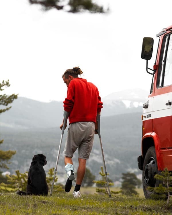 Max from @hummel_die_feuerwehr on crutches with a dog beside a red Mercedes-Benz 608D "Düdo" firetruck converted into a van, overlooking a scenic mountain landscape, representing the challenges of vanlife after an injury in a remote location on Crete.