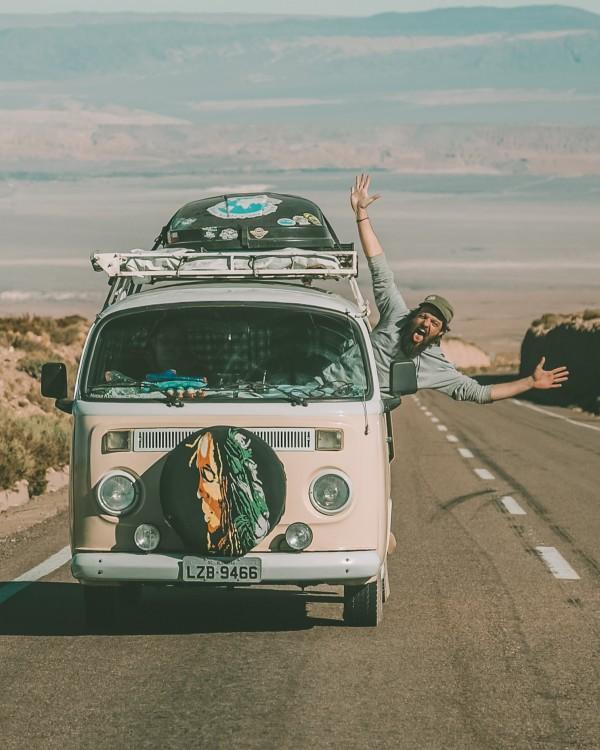 A person joyfully extends their arms out of the passenger window of a vintage Volkswagen van traveling on a desert road. The van, adorned with stickers and a front spare tire cover featuring a wolf design, conveys a sense of adventure against a backdrop of distant mountains under a clear blue sky.
