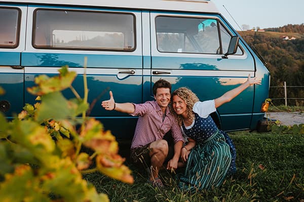 Thomas & Regina, from @edelweiss.on.the.road, pictured in the wine hills of Styria, Austria, wearing traditional Trachten clothing beside their VW T3 Vanagon "Edelweiss," capturing a moment at the beginning of their vanlife journey immersed in Austrian culture.