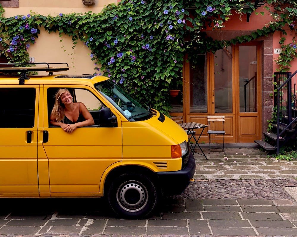 A joyful woman with blonde hair leans out of the driver's side window of a bright yellow van parked on a charming cobblestone street. Behind her, a vibrant climbing plant with purple flowers adorns the exterior of a classic European building with pastel-hued walls. The scene conveys a sense of travel and adventure in a quaint urban setting, with the woman's smile suggesting a carefree and exploratory spirit.