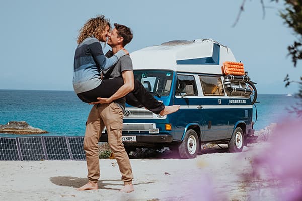 Thomas & Regina, from @edelweiss.on.the.road, are pictured on a beautiful beach in Lefkada, Greece. In this intimate moment, Thomas lifts Regina as they share a kiss, with their VW T3 Vanagon "Edelweiss" parked in the background near the turquoise waters. Solar panels are set up beside the van, highlighting their off-grid vanlife setup. The serene ocean and clear blue sky create a perfect backdrop, reflecting the freedom and romance of their vanlife adventure along the idyllic Greek coastline.