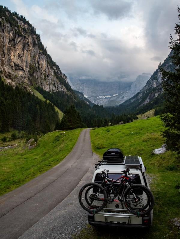 A white T4 van, with bicycles mounted on the back and a rooftop storage box, is parked on a narrow mountain road in a lush green valley. The van is surrounded by towering cliffs and dense forests, leading to mist-covered mountains in the distance. The scenic landscape captures the spirit of outdoor adventure and travel, reflecting the journeys shared by Christina and Ralf, known as @miles_together.