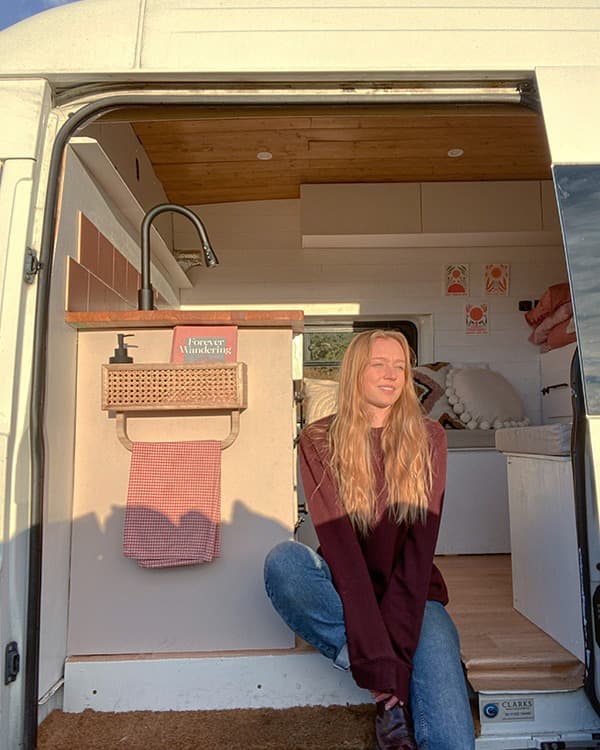 @tamzin_in_transit sitting in the doorway of her converted 2011 Ford Transit van, showcasing a cozy van life setup with a wood-paneled ceiling, a kitchenette with pink tiles, a farmhouse sink, and stylish decor. The open door reveals a bright and inviting interior, reflecting a minimalist and adventurous lifestyle.