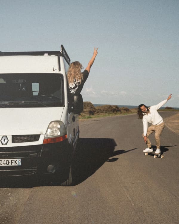 A young couple, @irisandmax, enjoying vanlife with their converted Renault Master van. The woman leans out of the van, raising a peace sign, while the man skateboards alongside on a scenic road with clear skies and nature in the background. Vanlife adventure captured in motion.