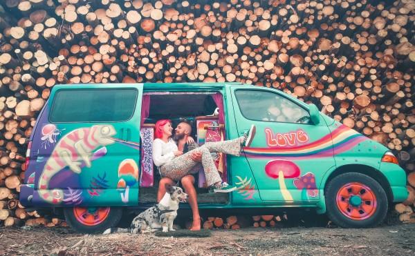 Joriën and Sandra from travelwildtobefree, sitting happily in their colorful, hippie-themed Toyota HiAce 4x4 camper van with their corgi Fudge, in front of a stacked pile of logs.