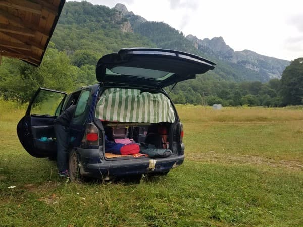 A Renault Espace microcamper parked in a lush green meadow, surrounded by mountains in Bulgaria. The back of the van is open, revealing a cozy, makeshift sleeping area with bedding and supplies. A person is seen arranging items inside the vehicle, preparing for a peaceful night in nature. The scene captures the essence of a simple and adventurous van life in a mountainous region.