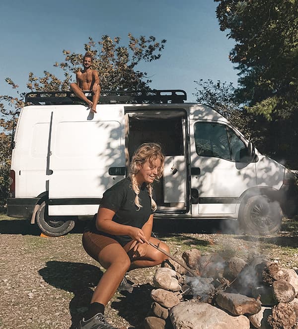 A young couple, @irisandmax, enjoying vanlife at a campsite with their converted Renault Master van. The woman tends to a campfire, while the man sits on top of the van, surrounded by trees and nature, capturing the essence of outdoor living and vanlife adventures.