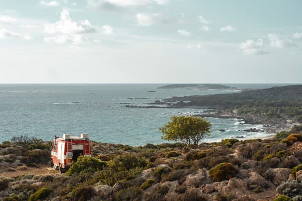 A red firetruck, @hummel_die_feuerwehr, converted into a camper, parked on a rocky coastline overlooking the sea, highlighting the vanlife experience in a remote coastal location on Crete’s south coast.