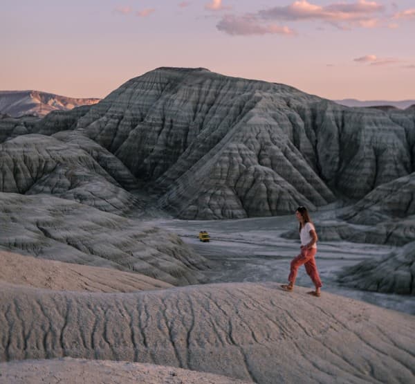 A woman, Astrid from @our_orbit_vanlife, in casual clothing walks on a rugged, rocky terrain at sunset in Nallıhan Ankara, Turkey. In the background, a yellow Mercedes Vario 816D camper van is parked among dramatic, eroded rock formations, highlighting the adventurous spirit and natural beauty of vanlife.