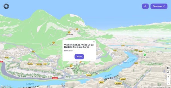 Interactive 3D map from Vanlifezone featuring the Via Ferrata Les Prises De La Bastille route in Grenoble, France. The map shows a detailed terrain of green mountains and the city's layout with buildings in relief, roads like D57 and D590, and the blue river L'Isère. A pop-up provides information on the Via Ferrata route, including the difficulty level, with a 'Route' button for navigation. Icons for Via Ferrata and a charging station are visible, aiding in adventure planning. The interface offers a 'Close map' feature and has Mapbox and OpenStreetMap attributions.