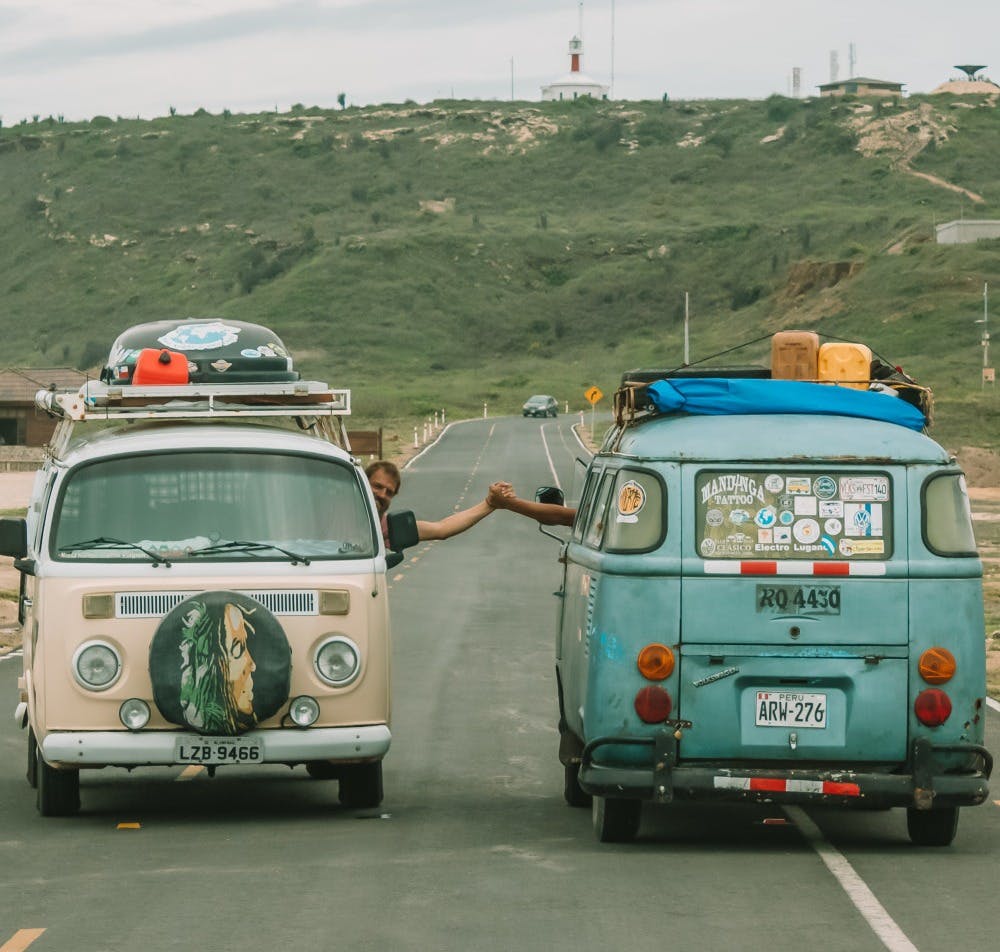 Two vintage Volkswagen vans parked on a paved road, with their drivers reaching out for a handshake between the vehicles. One van is cream-colored with a large artistic face painting on the spare tire cover, while the other is blue with a collection of travel stickers and a surfboard on the roof. In the background, there's a hilly landscape with a lighthouse, under an overcast sky, suggesting a spirit of camaraderie and travel adventure.