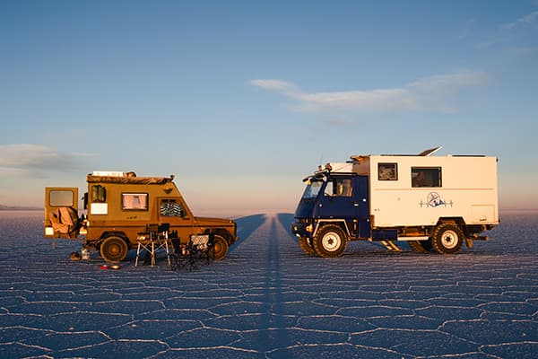 Two overland camper vehicles, a green Puch 230GE and a large blue and white expedition truck, parked facing each other on the vast salt flats of Salar de Uyuni at sunset.