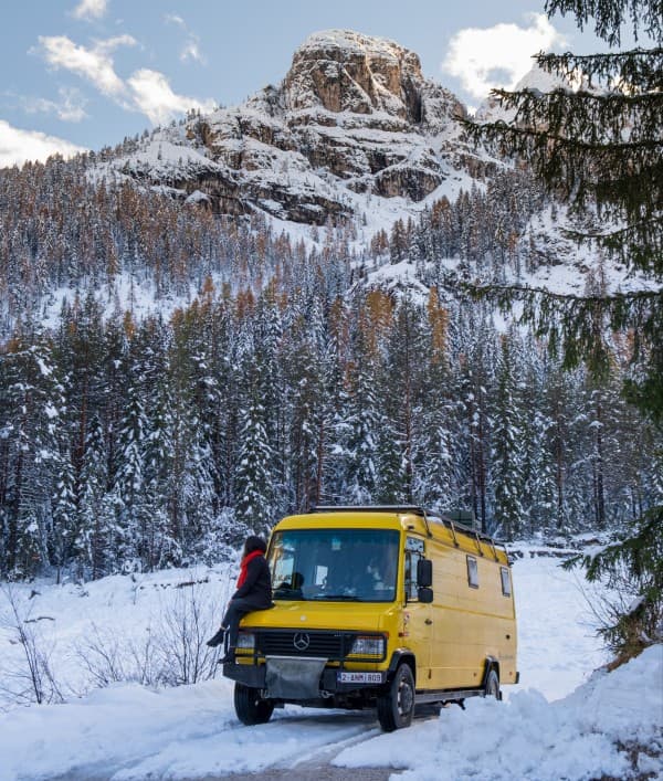 Yellow Mercedes Vario 816D camper van from @our_orbit_vanlife parked in a snowy mountainous landscape with pine trees and a rocky peak in the background. One person, dressed in a red scarf and dark winter clothing, sits on the front bumper of the van, enjoying the serene winter scenery. The scene captures the essence of vanlife during winter, highlighting the adventure and beauty of nature.