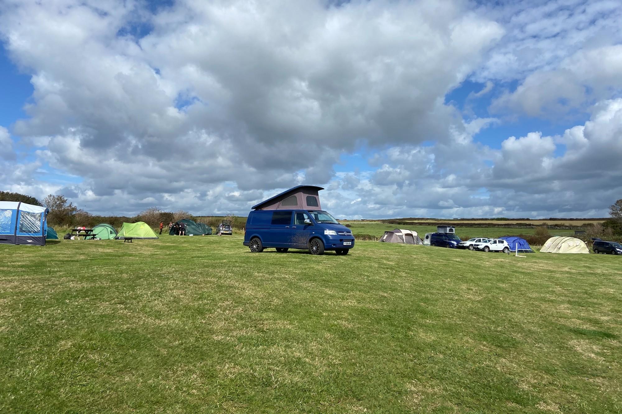 A blue Volkswagen T5 camper van with pop-up roof parked on a grassy field at a campsite in the UK, under a sky with dramatic clouds.