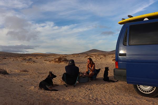 Jo and Georg from triptovantasia, with their dogs, sitting beside their blue VW T4 van in a desert landscape on the Canary Islands, enjoying a meal during their vanlife journey.