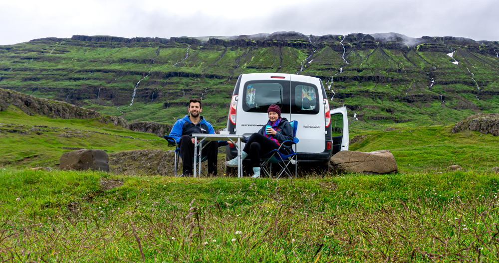 A couple enjoying a meal at a portable table with chairs next to a white Renault Kangoo van, parked in the Icelandic countryside with cascading waterfalls in the distance, lush green hills, and overcast skies.