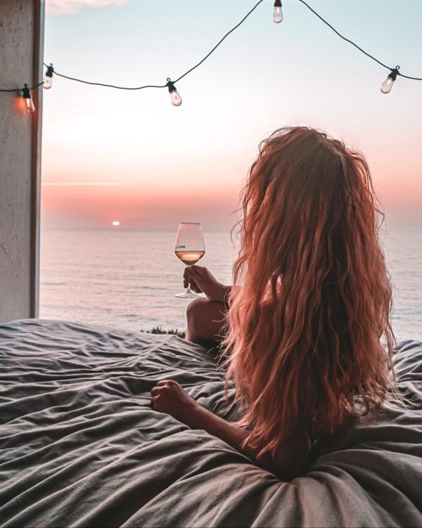 A blond woman enjoying a sunset view over the ocean from the back of a converted box truck camper, with string lights and a glass of wine.