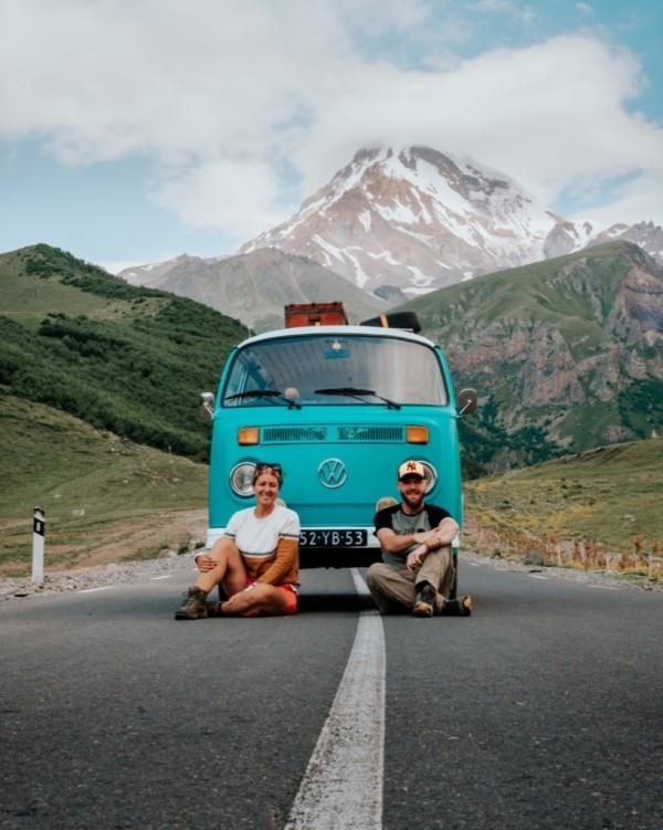 A man and a woman, Milene and Yuri from @mygrations.nl sitting on the road in front of a turquoise Volkswagen T2 van with a mountainous landscape in the background. The van is parked on a clear road with the scenic snow-capped peaks of the Greater Caucasus Mountains, possibly near Mazeri, Georgia, in the distance. The bright blue sky with scattered clouds enhances the adventurous spirit of their journey along the ancient Silk Road. The image captures the essence of van life and road trips through remote and beautiful landscapes.