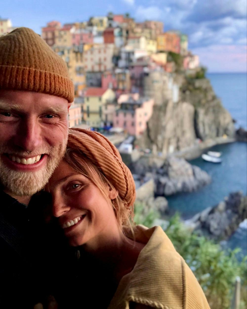 A close-up selfie of a smiling couple, with the man wearing a warm beanie and the woman sporting a headscarf, both enjoying a cozy moment. They are positioned against a blurred backdrop of Manarola, Cinque Terre, with its distinctive colorful houses perched on rugged cliffs beside the sea. The image captures the warmth of the couple's relationship set against the romantic and picturesque Italian Riviera, at what appears to be dusk, adding a soft, glowing light to the scene.