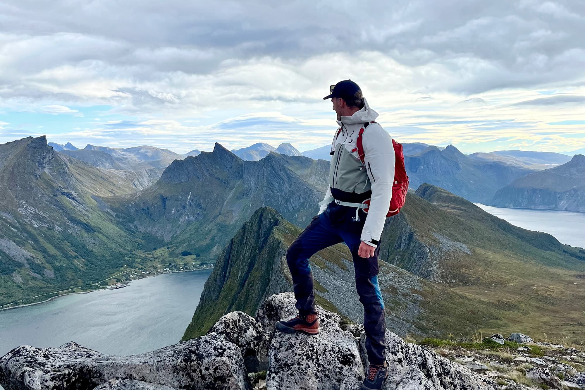 Chris (@van4life__) standing on a rocky peak during a hike in Norway, overlooking stunning mountain ranges and a fjord. The image captures the expansive natural beauty of Norway's dramatic landscapes, with Chris in outdoor gear and a red backpack, embodying the spirit of adventure and exploration.