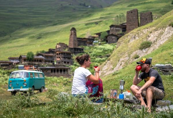 Milene and Yuri enjoy a coffee break outdoors in a scenic mountain setting with their turquoise Volkswagen T2 van, Alexine, parked nearby. The background features the historic village Dartlo in Georgia with stone watchtowers and traditional buildings set among rolling green hills, reflecting a peaceful moment of van life and cultural exploration on their Silk Road journey.