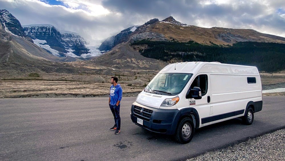 A man standing by a Ram Promaster 159 Extended van parked on the Icefield Parkway with dramatic overcast skies above and rugged mountain scenery in the background, conveying the majestic and wild nature of the Canadian Rockies.