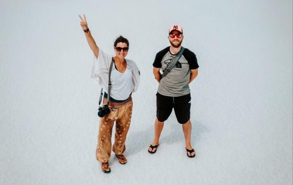 Milene and Yuri from @mygrations.nl standing on a vast salt flat, with Milene making a peace sign and holding a camera, and Yuri standing beside her, both wearing casual travel clothing. The white, expansive landscape creates a minimalist backdrop, highlighting their adventurous spirit as they explore unique destinations on their journey along the Silk Road. The image captures a moment of travel, adventure, and cultural exploration.