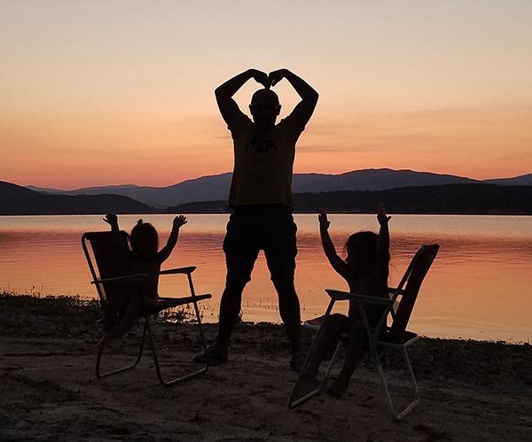 A silhouette of a man and two young girls enjoying the sunset by a serene lake during their van life adventure in Bulgaria. The man stands in the middle, forming a heart shape with his hands above his head, while the girls sit in camping chairs on either side, raising their arms joyfully. The scene captures a peaceful and loving moment at the end of a day spent in nature.