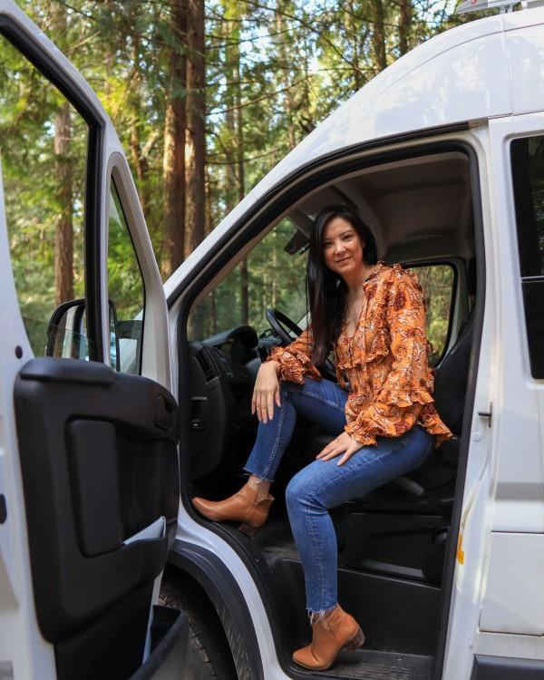 Emily Jay from @emsvanlife sitting in the driver's seat of a RAM van in a forest setting, representing her vanlife journey. She is wearing a patterned orange blouse and jeans, with trees visible in the background, illustrating her outdoor lifestyle.