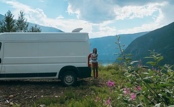 A person stands next to a white van parked amidst a wildflower-strewn landscape with a mountainous backdrop under a dynamic sky. The van's roof features a vent, illustrating the allure of mobile living and exploration in a serene, natural setting, perfect for outdoor and travel enthusiasts.