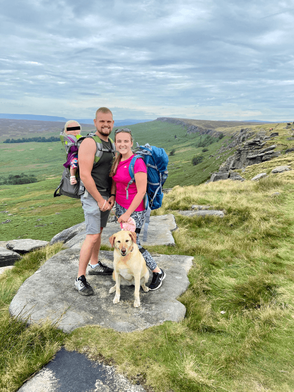 A smiling family of three with a baby in a carrier and a golden retriever dog, standing on a rocky outcrop during a hike in the UK countryside, with rolling hills in the background.