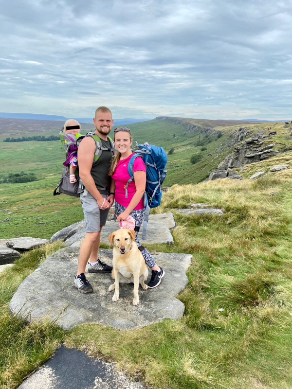 A smiling family of three with a baby in a carrier and a golden retriever dog, standing on a rocky outcrop during a hike in the UK countryside, with rolling hills in the background.