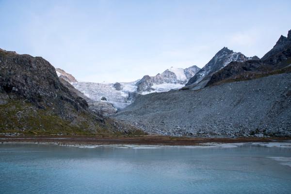 A stunning view of a glacier in the Valais region of Switzerland, with rocky mountains and snow-capped peaks in the background. In the foreground, a serene glacial lake reflects the surrounding landscape under a clear sky. This image captures the natural beauty and rugged terrain of the Swiss Alps, showcasing the breathtaking scenery that Christina and Ralf from @miles_together explore during their adventures.