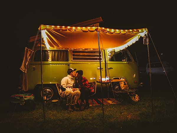 A couple, Anna and Christoph from @meetheinrich, enjoying a cozy evening outside their vintage VW T2 bus, Heinrich, under a lit awning during a night camping adventure. The warm glow of string lights adds to the relaxing atmosphere of their vanlife experience.