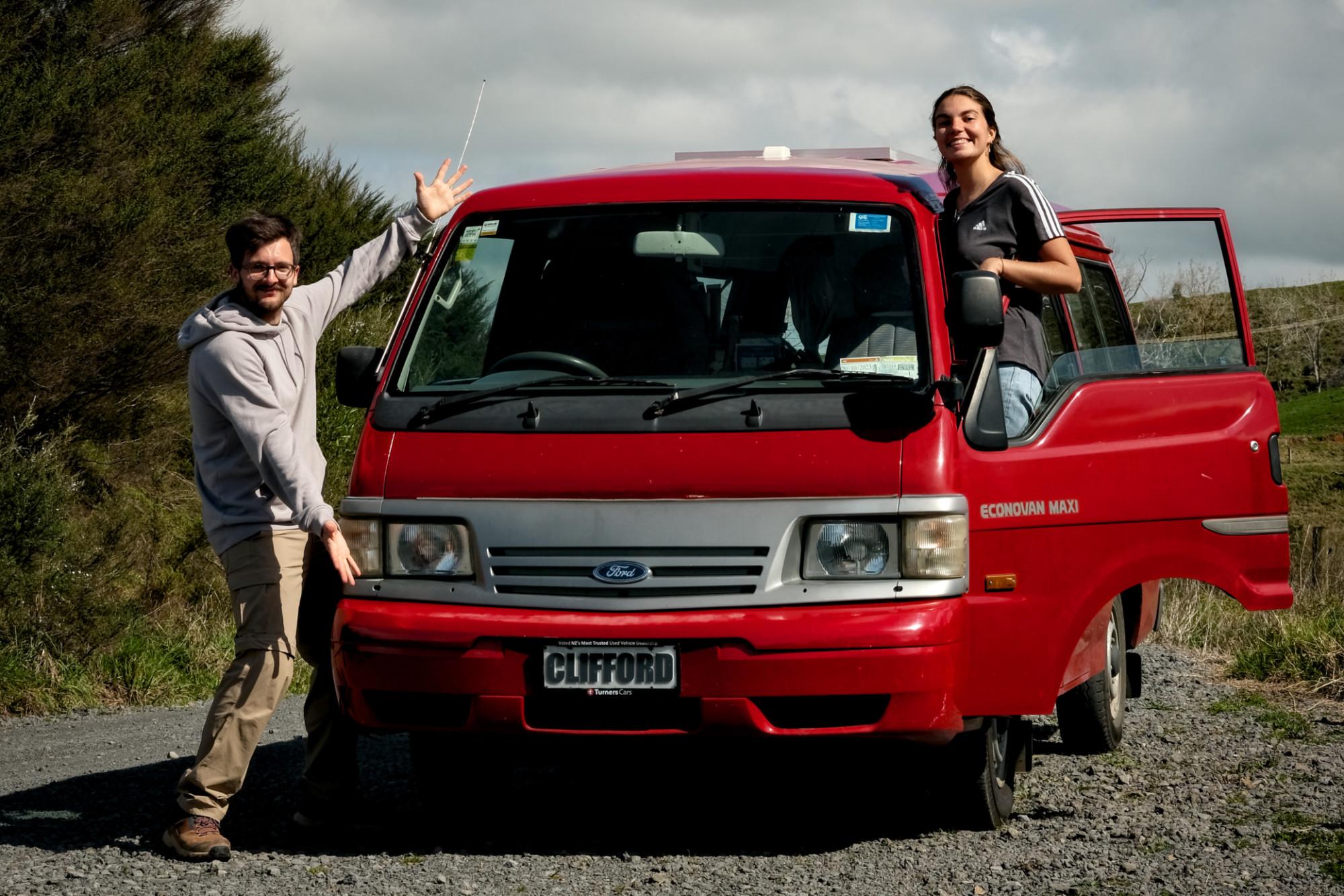 David and Claire posing with their red Ford Econovan named Clifford, parked on a gravel path amidst lush greenery in New Zealand. David gestures enthusiastically towards the van while Claire leans out of the passenger door, showcasing their adventurous vanlife journey. This image captures the spirit of exploration and travel photography by the authors, @carnet_declaireurs.