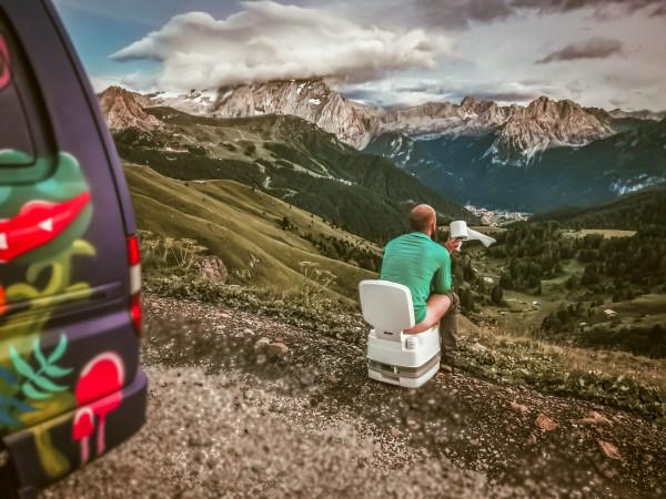 Joriën from travelwildtobefree, sitting on a portable toilet with a roll of toilet paper, enjoying a scenic mountain view from their colorful camper van.