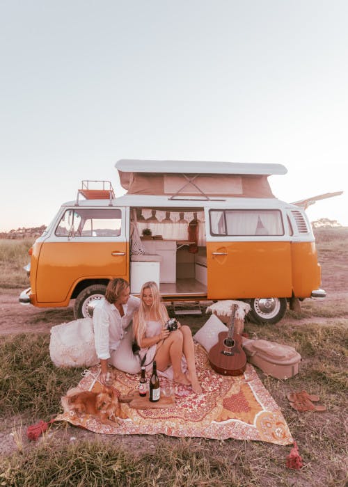 A couple enjoys a romantic picnic at dusk next to a classic yellow and white camper van with the pop-top open. They are sitting on a colorful rug adorned with pillows and a guitar, surrounded by peaceful countryside. The woman, holding a vintage camera, is listening to the man as he speaks, while a small, fluffy dog rests beside them. A bottle of wine and two glasses complete the intimate setting under a soft, pastel sky. @75vibes_ 09/2020 - Vanlifezone (Issue 1)