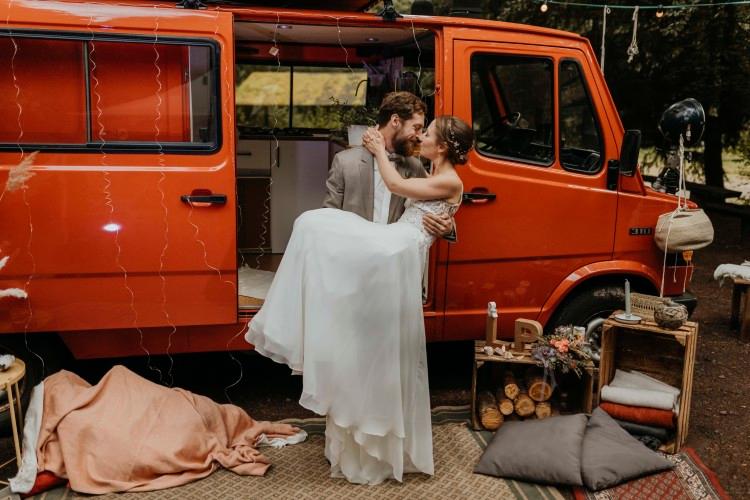 A newlywed couple sharing a tender moment beside a vibrant orange vintage van. The bride, in a flowing white gown, and the groom, in a smart grey suit, are embracing and gazing into each other’s eyes. The van’s door is open, revealing a cozy interior, and the scene is adorned with rustic decorations including a string of lights, blankets, pillows, and a makeshift wooden shelf with various personal items, set in a tranquil outdoor setting with trees in the background. @the.redcamper 10/2020 - Vanlifezone (Issue 2)