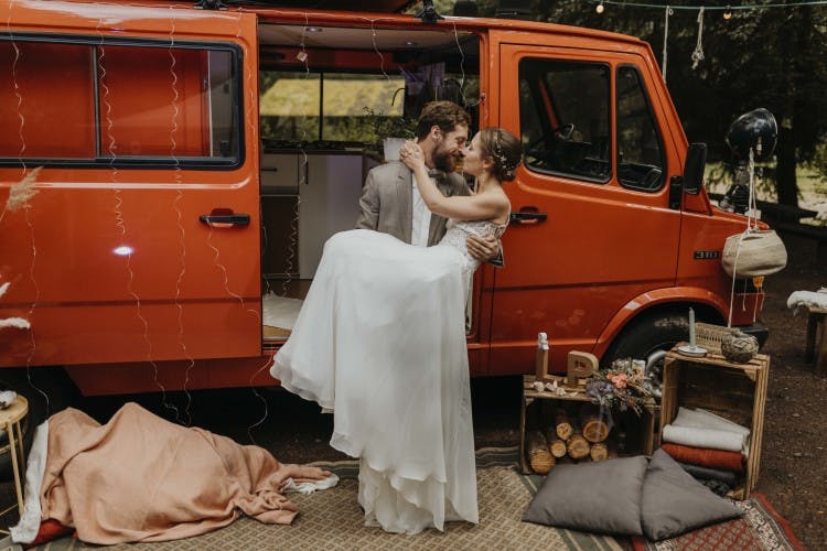 A newlywed couple sharing a tender moment beside a vibrant orange vintage van. The bride, in a flowing white gown, and the groom, in a smart grey suit, are embracing and gazing into each other’s eyes. The van’s door is open, revealing a cozy interior, and the scene is adorned with rustic decorations including a string of lights, blankets, pillows, and a makeshift wooden shelf with various personal items, set in a tranquil outdoor setting with trees in the background. @the.redcamper 10/2020 - Vanlifezone (Issue 2)
