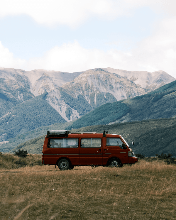 Red camper van parked in a scenic mountainous area of New Zealand, showcasing the vanlife experience. The vehicle is set against a backdrop of lush green hills and rugged mountains under a partly cloudy sky, emphasizing the adventurous spirit of travel photographers exploring the natural beauty of New Zealand.